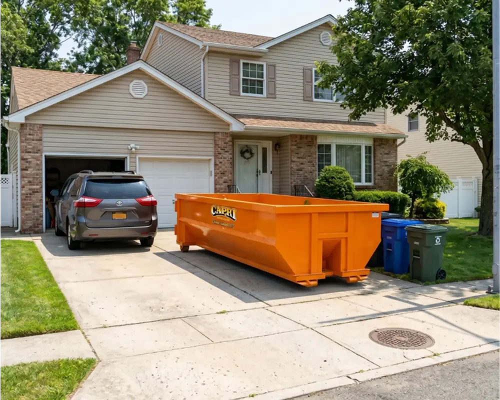 A bright orange 10-yard roll-off dumpster branded with the "Capri" logo sits on a concrete residential driveway. In the background is a two-story tan house with brick accents, a gray minivan parked in the open garage, and blue and green trash bins nearby.